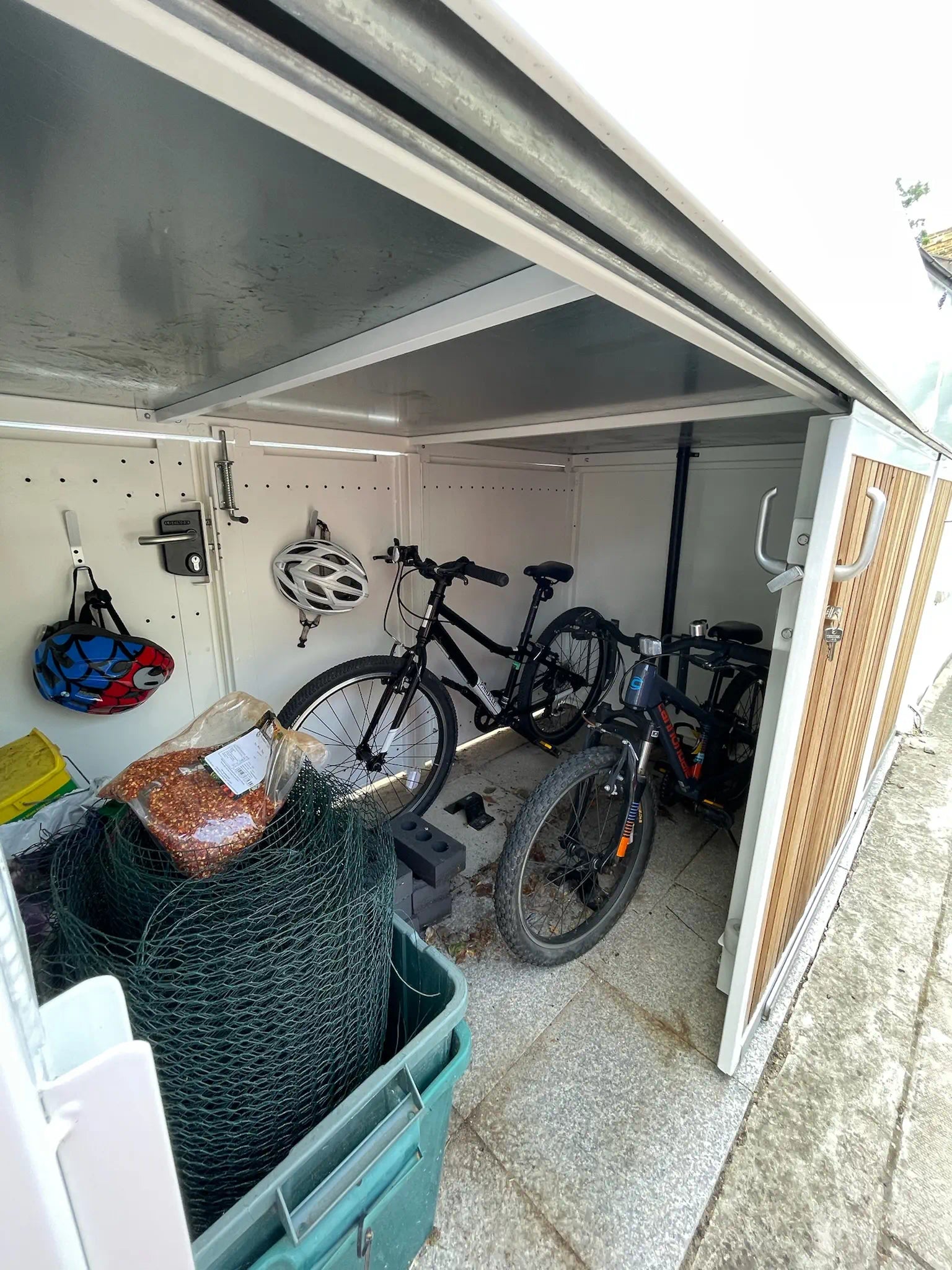 Open sliding-door bike store beside a bay window, showing bicycles, helmets, and garden tools neatly stored inside.