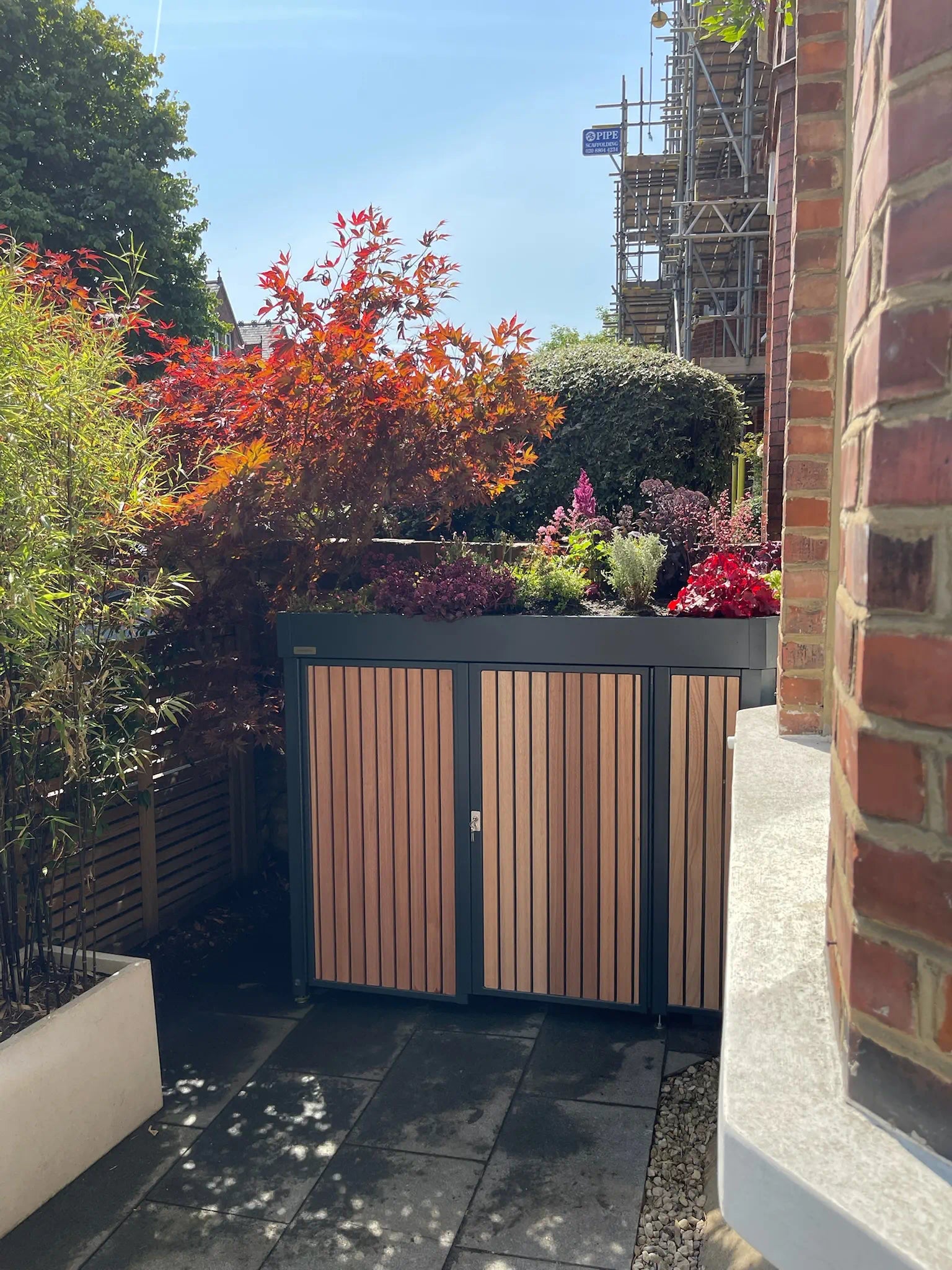 Bespoke Verde Forma bike store viewed from house entrance, wrapping around bay window with biodiverse green roof, timber cladding, and secure locking system in London front garden.