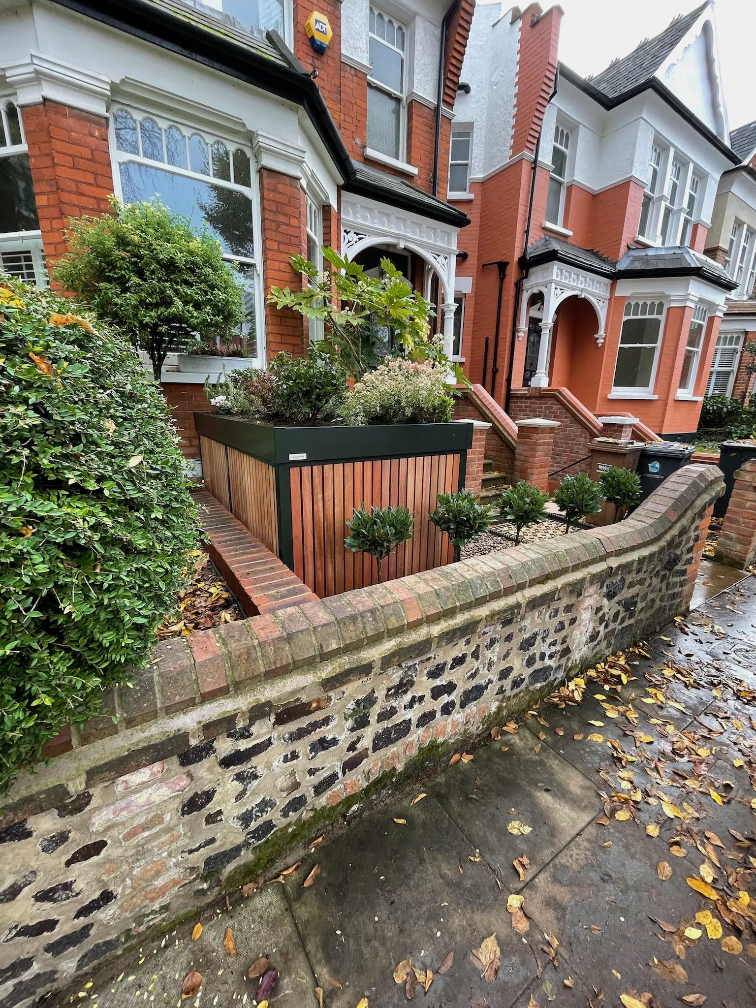 Street view from the left of deep green front garden bike store with timber cladding, blending into a London terraced street