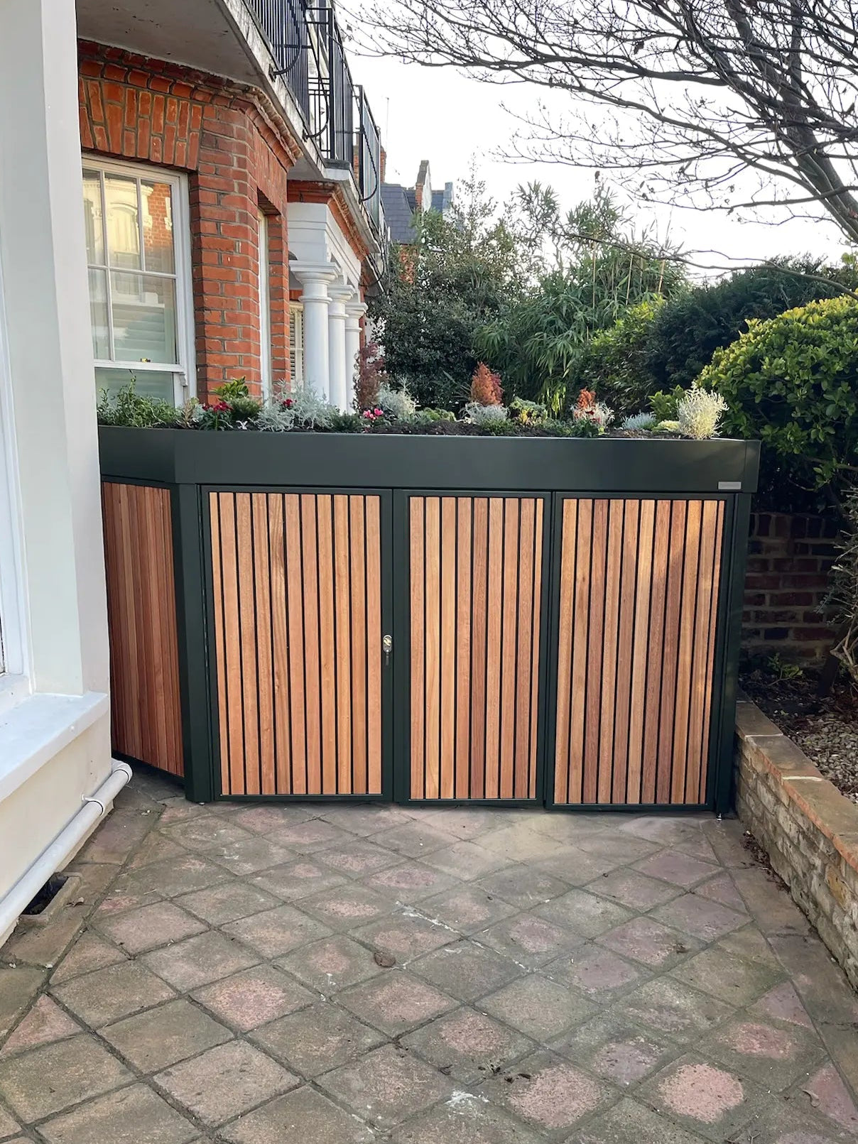 Front view of deep green side bike store with closed doors and planted roof, discreetly integrated into a London conservation area