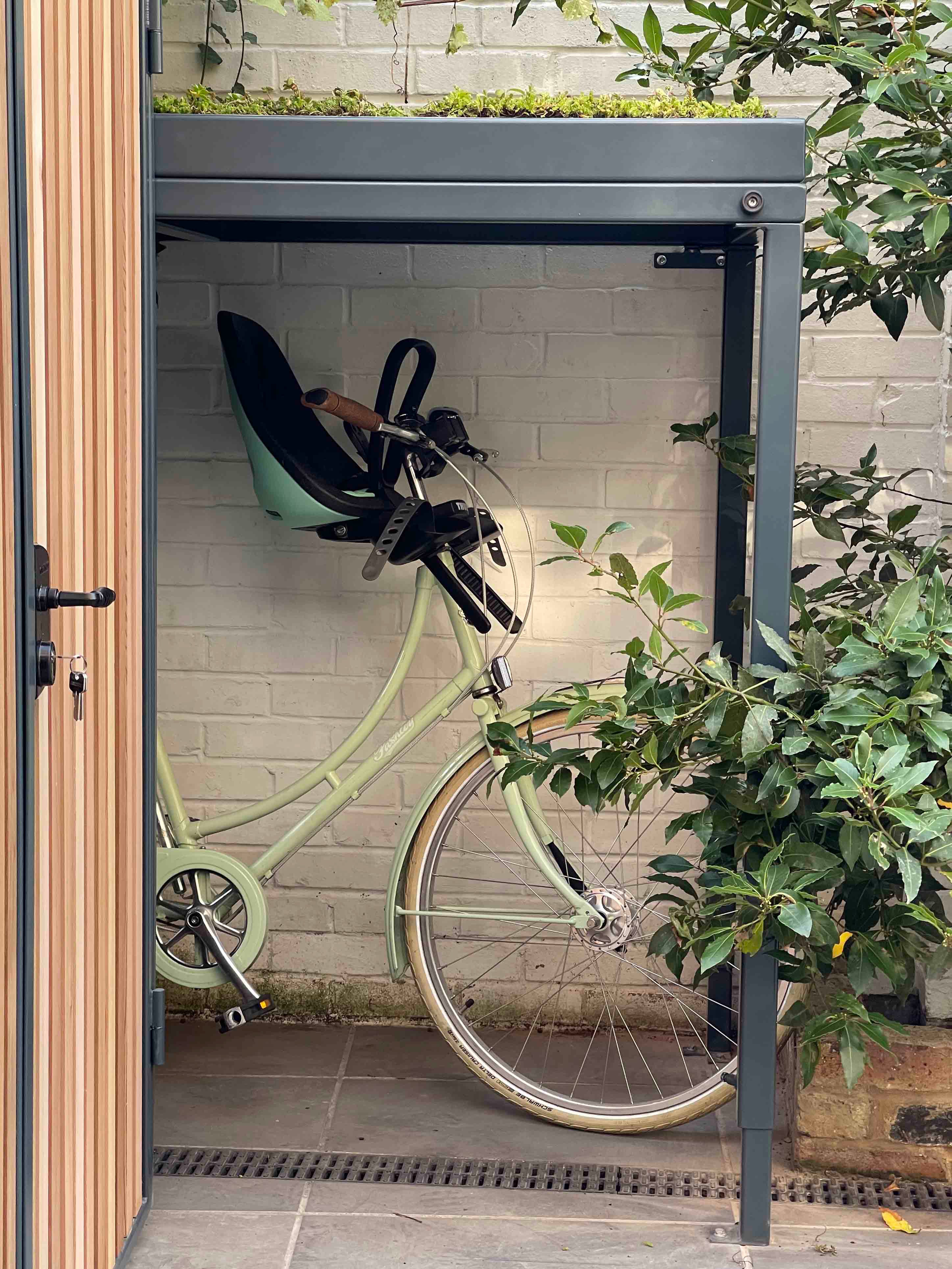 Detail view of bike shelter protecting bike with child seat on, with extensive green roof above 