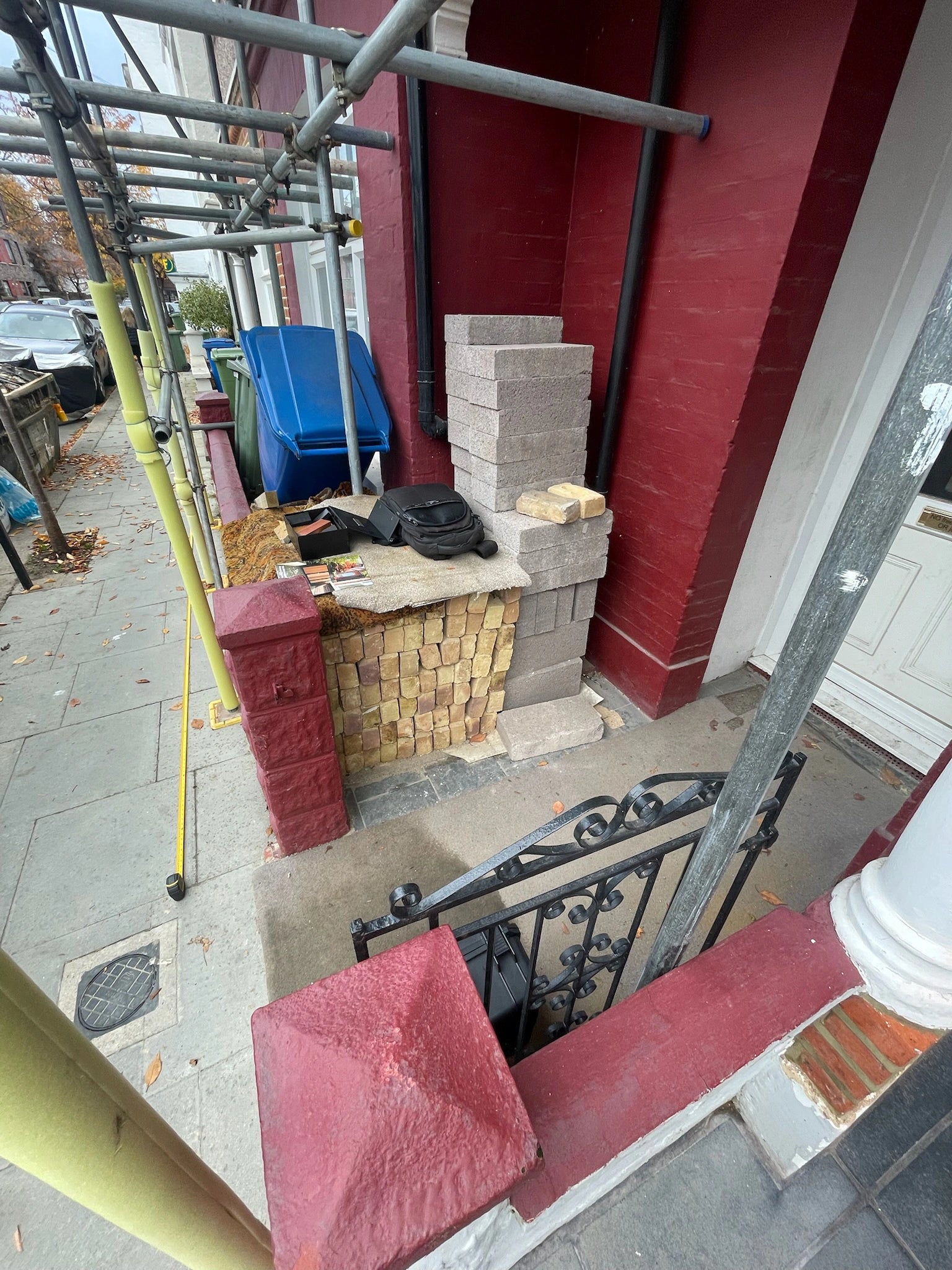Before: front entrance of a London terrace under scaffolding, with building materials and bins staged beside the boundary wall ahead of a front garden bike and bin store.