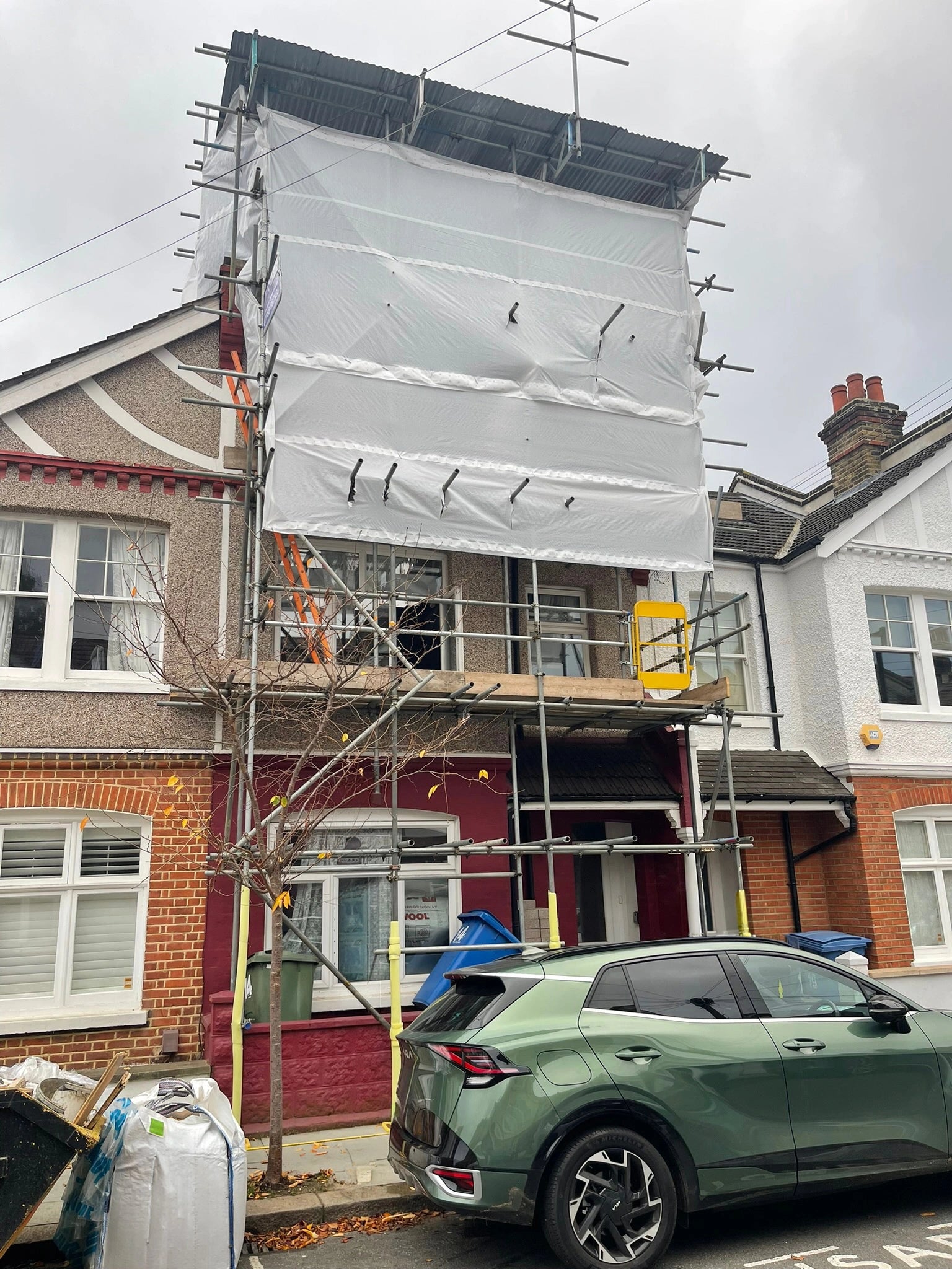 Before: full elevation of a London terrace covered in scaffolding and protective sheeting during renovation, ahead of a front garden bike and bin store.