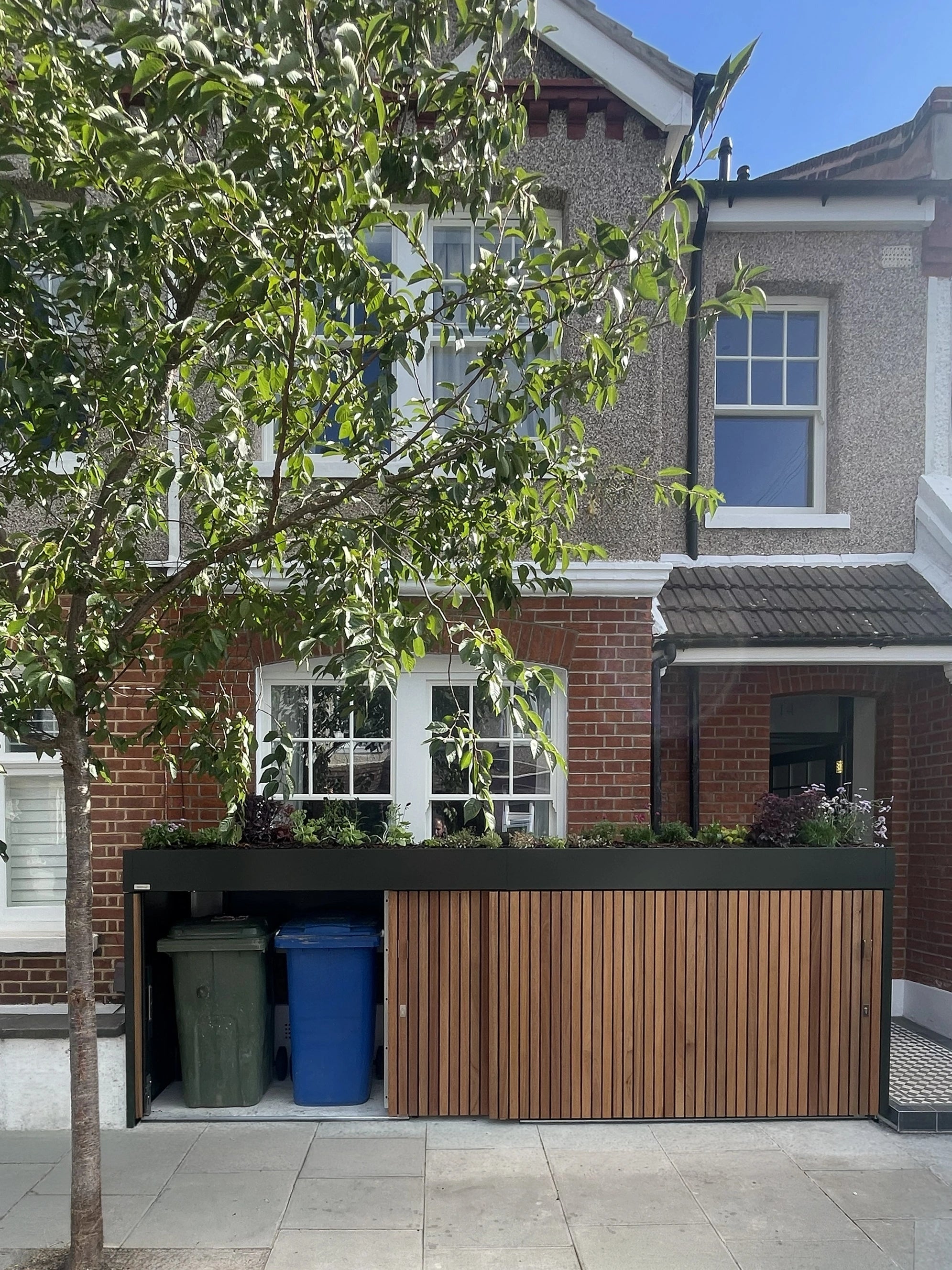 Front garden bike and bin store with doors open, showing the family's green and blue wheelie bins concealed beneath a planted roof.