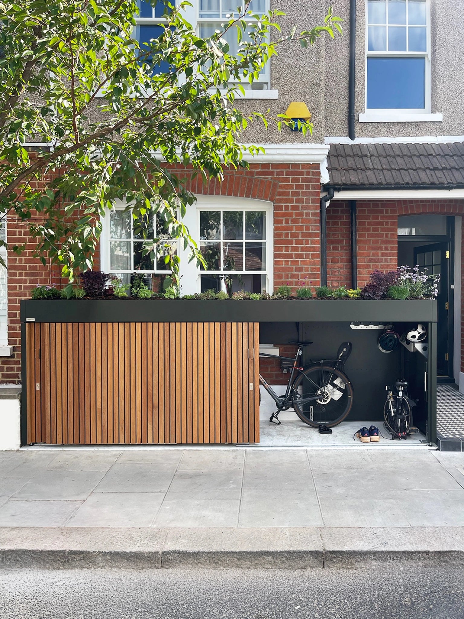 Front garden bike and bin store with doors open, showing bikes and helmets stored beneath a planted roof in a London terrace garden.