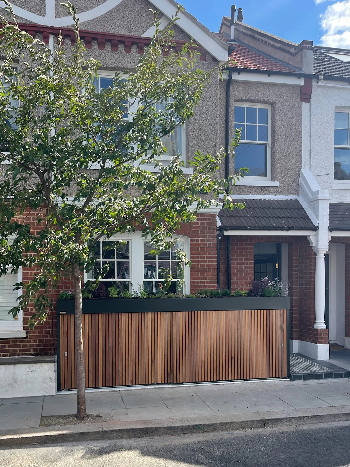 Front garden bike and bin store with closed timber doors, set against a London terrace house and a young tree.