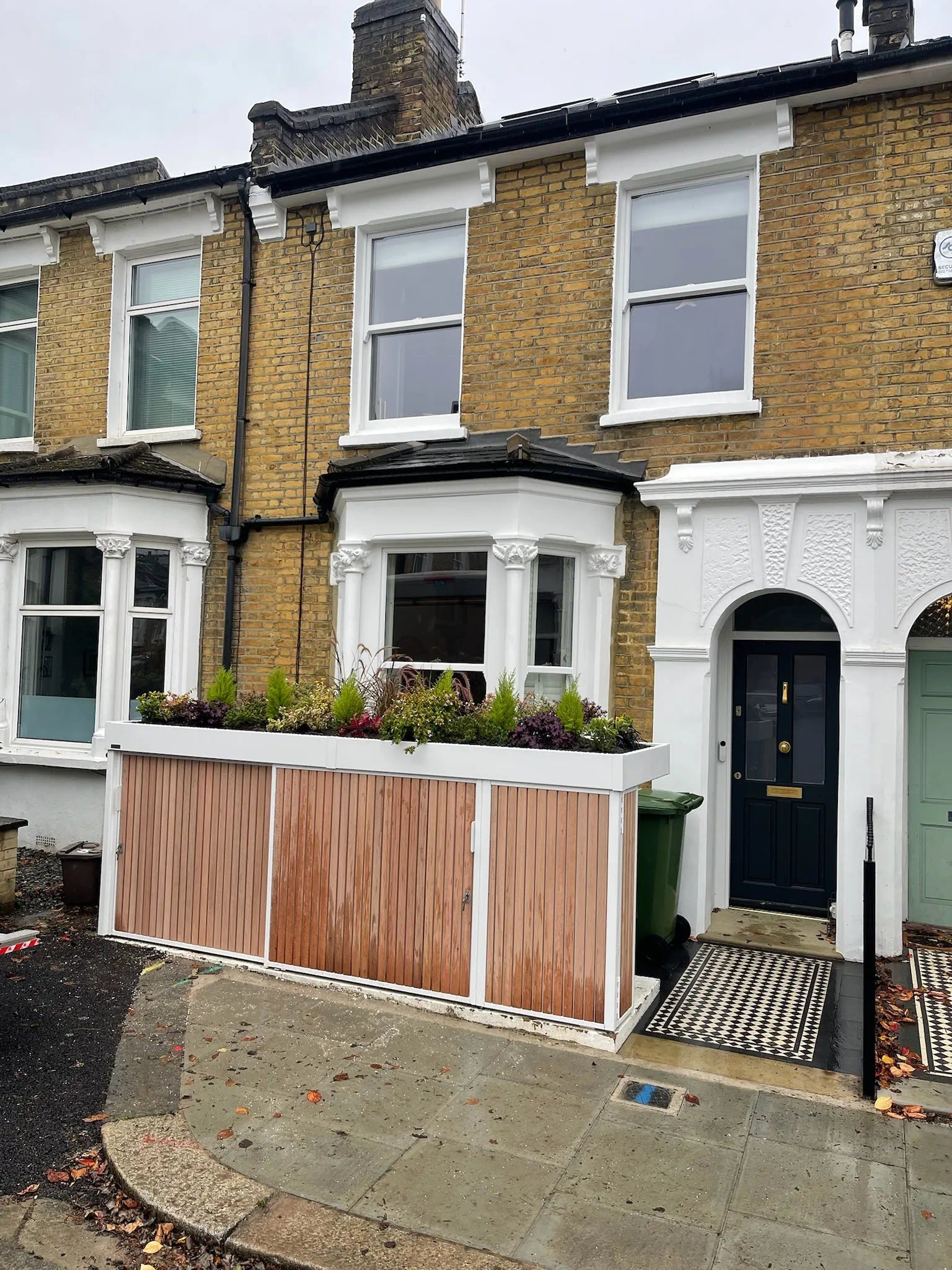 Compact linear bike store with white frame, hardwood cladding and planted roof replacing front garden wall.