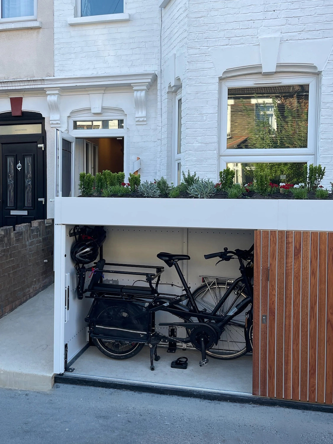 Front Garden store built into a replaced front wall of a white-painted London terrace, doors open to show a cargo bike, bike and helmet stored beneath a planted roof. 