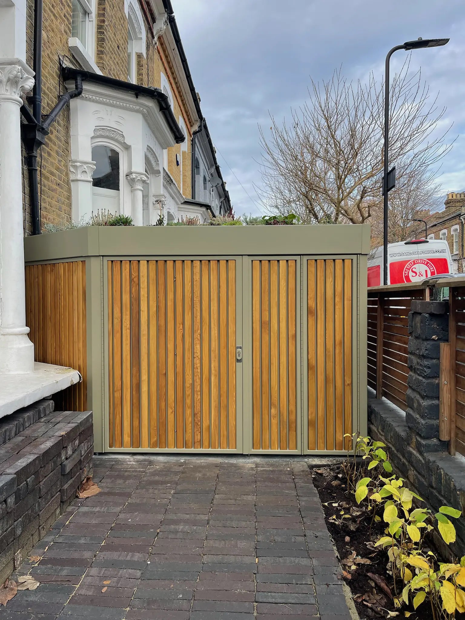 Close-up of olive-grey metal cycle store with walnut timber cladding in London front garden